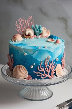 a blue cake decorated with sea shells and seashells is on a glass platter