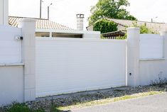 a white fence is in front of a house on the side of a road with grass and bushes