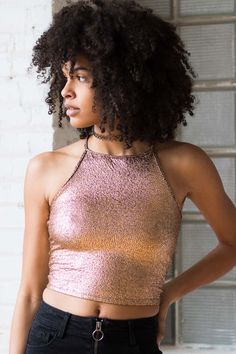 a woman with curly hair wearing a pink top and black jeans standing in front of a brick wall