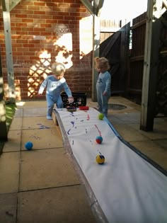 two young boys playing with balls on an outdoor play table in front of a brick building