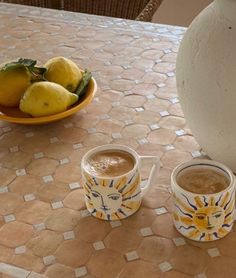 two cups of coffee sit on a table with lemons and a bowl of fruit