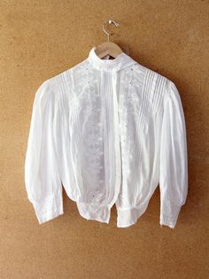 This is an antique Edwardian white blouse with pin tuck detailing. The delicate sheer cotton top features emboss embroidered flowers and lace accented sleeves. It has a button closures and hook closure at the neck.  CONDITION In good condition for age.  There is a small hole on the right side and on two on the right sleeve, a small old repair stitch on the left shoulder and bottom front, and it is missing the original hook closure at neck.  SIZE & MEASUREMENTS Approximate FitMedium Bust45 inch Classic White Lace Tops, Victorian White Cotton Top, Victorian Style White Cotton Top, Classic White Broderie Anglaise Tops, Classic White Tops With Broderie Anglaise, Classic White Tops With Lace Work, White Victorian Style Top With Lace Work, Classic Formal Tops With Floral Embroidery, Fitted Classic Tops With Broderie Anglaise