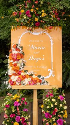 a sign with flowers on it sitting in front of some trees and bushes at a wedding