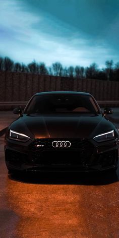 the front end of a black car parked in a parking lot at night with its lights on