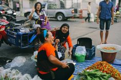 three women sitting on the ground in front of various food items and people standing around