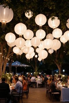 people are sitting at tables with white lanterns hanging from the ceiling above them and trees in the background