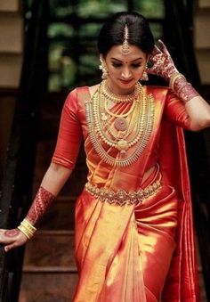 a woman in an orange and yellow sari walking down stairs with her hands on her head