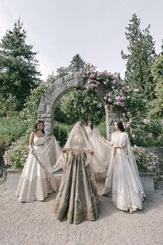 three women dressed in wedding gowns and veils standing under an arch with flowers on it