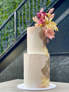 a three tiered white cake with gold leaf designs and flowers on the bottom layer