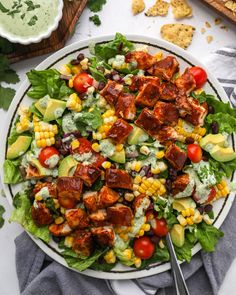 a salad with meat, avocado, tomatoes and corn on it in a white bowl