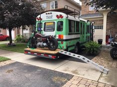 a green bus with a motorcycle on the back of it's trailer in front of a house