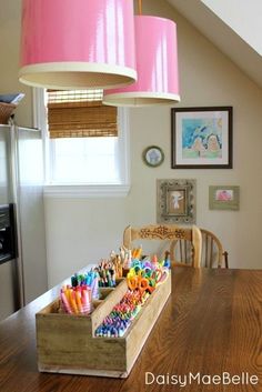 a pink lamp hanging over a wooden table with lots of pens and pencils in it