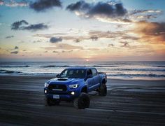 a blue truck driving on top of a sandy beach next to the ocean at sunset