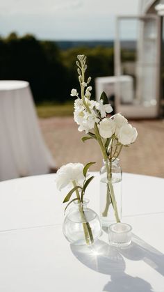 two clear vases with white flowers are sitting on a round table in front of an outdoor setting