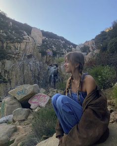 a woman sitting on top of a large rock next to a lush green hillside covered in rocks