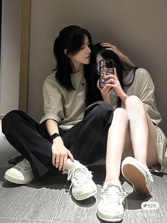 two women sitting on the floor taking pictures with their cell phones and one is wearing white sneakers