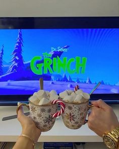 two people holding up mugs with marshmallows in front of a tv