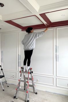 a woman on a ladder painting the ceiling in an empty room with white walls and red trim