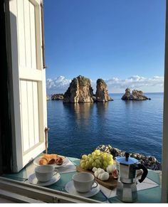 an open window overlooking the ocean with fruit and coffee cups on a table in front of it