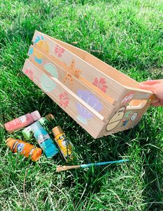 a hand holding a wooden box filled with crayons on top of green grass