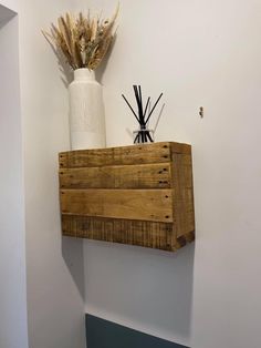 a white vase with dried flowers on top of a wooden shelf above a toilet paper dispenser