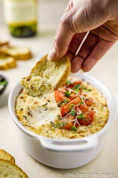 a hand dipping a piece of bread into a bowl of dip with tomatoes and cheese