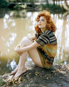 a woman with red hair is sitting on the bank of a river and looking off into the distance