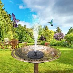 a bird bath in the middle of a garden with two birds flying over it and another bird on the other side