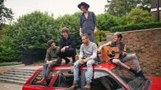 four men are sitting on top of a red car and one man is playing the guitar