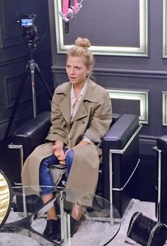 a woman in a trench coat sitting on a chair with a microphone and camera behind her