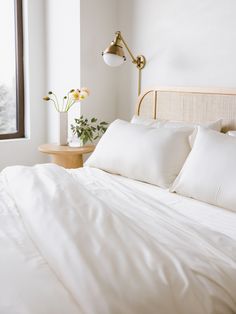 a bed with white sheets and pillows next to a lamp on a table in front of a window