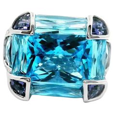 A bold and captivating ring featuring a cushion-cut blue topaz as the centerpiece, weighing 11.0 carats. The vibrant blue hue of the topaz is complemented by fancy-cut iolites, totaling 2.62 carats, in a light purple color. Crafted in 18k white gold, the ring showcases a contemporary design with a robust setting that includes diamond accents. This fashion ring is substantial yet elegant, weighing 13.8 grams. Ideal for making a statement with any outfit. Ring Size 7.25 Weight: 13.8 grams Bellari Ring, Three Stone Emerald Cut Ring, Emerald Cut Ring, Swiss Blue Topaz Ring, Fun Bracelet, Emerald Cut Rings, Blue Sapphire Diamond, Gold Band Ring, Modern Ring