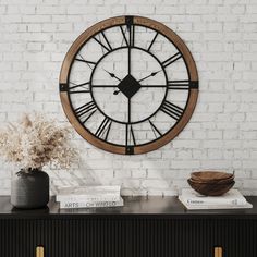 a large clock mounted to the side of a white brick wall next to a vase with dried flowers