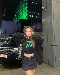 a woman standing in front of a car wearing a black jacket and skirt with her hands on her head