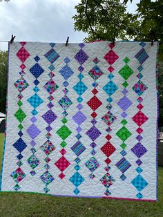 a colorful quilt hanging on a clothesline in the grass with trees and sky in the background