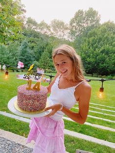 a woman holding a cake with candles on it in front of a lawn and trees