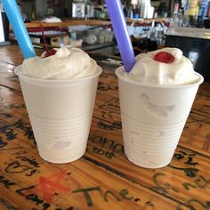 two drinks with whipped cream and cherries in plastic cups sitting on a wooden table