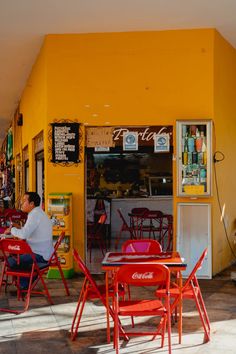 a man sitting at an orange table in front of a yellow building with red chairs