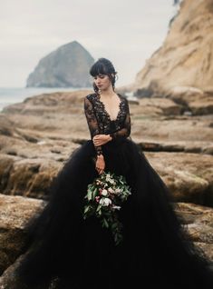 a woman in a black dress standing on rocks near the ocean wearing a flower crown