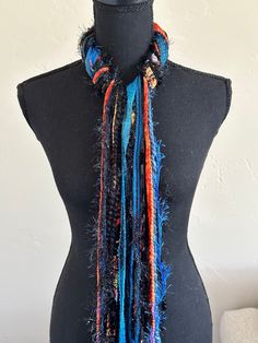 "This bright and colorful handmade scarf consists of blues, oranges, and black including a colorful hand sewn textile ribbon for added fun.  I utilize a variety of yarns in each of my scarves and no two are ever the same.  Each scarf is made up of a selection of Sari Silk, Eyelash, Ribbon, Ladder, Fuzzy, Sparkly, Pouf Pom Pom, Boucle, Flag Ribbon, Nub, Pluscious, Hand Spun, and Brushbound. It is approximately 72\" long. This is an open fringe style, tied at the middle. It is a medium weight scarf. Check out this YouTube link https://www.youtube.com/watch?v=5LYAEz777AU for a demonstration of the many ways that you can style this scarf." Blue Festival Scarf, Artsy Multicolor Handmade Scarves, Artsy Handmade Multicolor Scarves, Bohemian Black Handmade Scarves, Blue Bohemian Handmade Scarves, Blue Handmade Bohemian Scarves, Bohemian Black Handmade Scarf, Handmade Blue Yarn Scarf, Teal Scarf