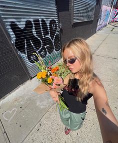 a woman is holding flowers on the sidewalk in front of a graffiti - covered building