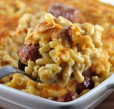 a spoon full of macaroni and cheese with sausage on it, in a white casserole dish