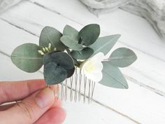 a hand holding a comb with flowers and leaves attached to it on a white wooden surface