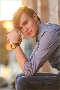 a young man sitting on top of a wooden bench next to a building and holding his hand near his face