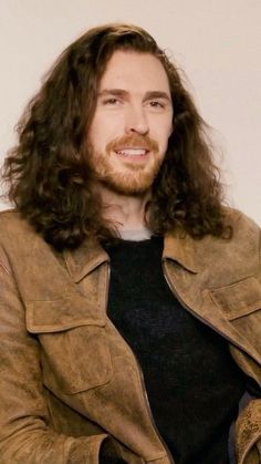 a man with long hair wearing a brown leather jacket and black shirt sitting in front of a white wall