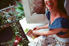 a woman sitting on the ground next to some flowers and potted plants in front of a window