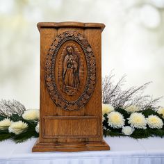 a wooden statue on top of a table with white flowers in the foreground and an image of virgin mary above it