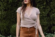 a woman standing in front of a bush wearing brown pants and a tan top with a tie around her waist