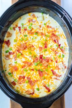 a crock pot filled with cheese, bacon and green onions on top of a wooden cutting board