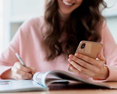 a woman sitting at a table looking at her cell phone and writing in a notebook